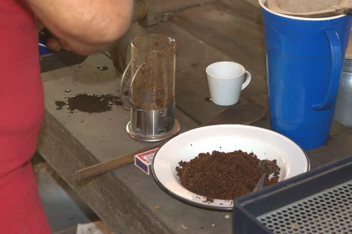 Prato esmaltado com o café moído na hora e uma colher, ao lado da prensa ainda suja do pó, sobre a bancada de madeira