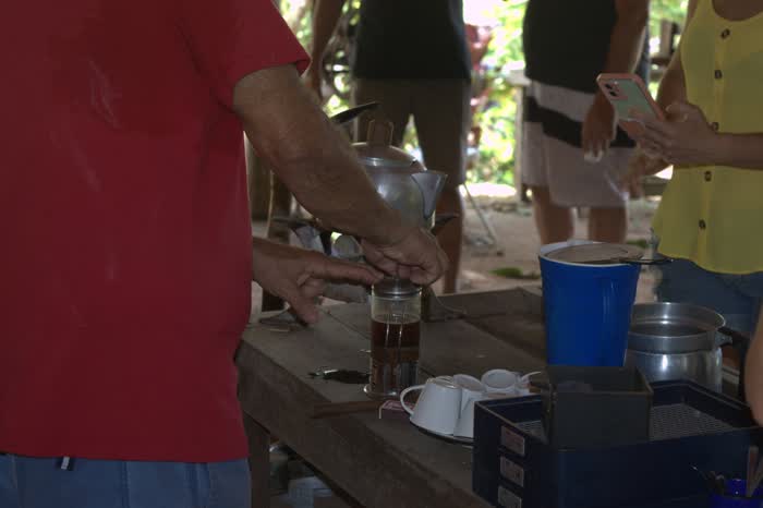 Mãos apertando o êmbolo da prensa cheia de café recém-coado, com as xícaras empilhadas e a chaleira de ágata ao lado