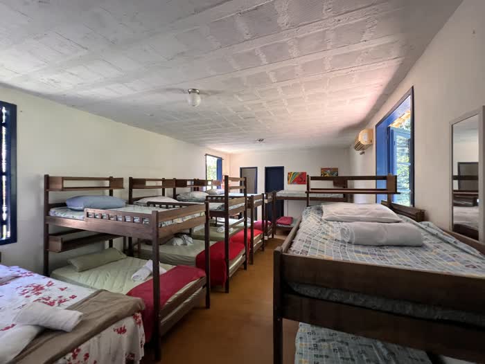 Alojamento comprido com várias beliches de madeira em duas fileiras, roupa de cama variada e janelas azuis nas duas pontas