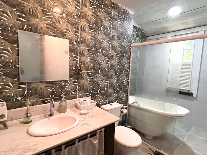 Banheiro com parede de azulejos estampados de folhas de costela-de-adão, pia de mármore com espelho e banheira com box de vidro