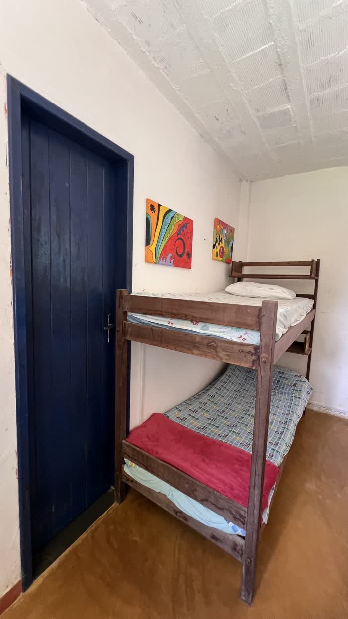 Beliche de madeira encostado na parede branca ao lado de uma porta azul, com cobertor vermelho na cama de baixo e dois quadros coloridos na parede