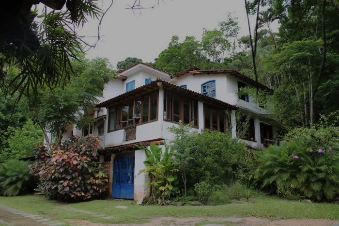 Fachada da casa-sede branca, de janelas azuis e telhado de telha vermelha, cercada pelo jardim e pela mata