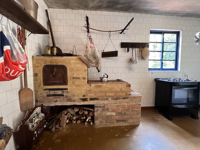 Cozinha da roça com fogão a lenha de tijolo e alvenaria, chaleira sobre a chapa, lenha empilhada embaixo e uma placa de madeira escrita “Sabor da Roça” na parede de azulejo branco
