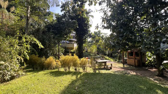 Jardim gramado com quiosque de madeira e mesa de pedra, à sombra das árvores