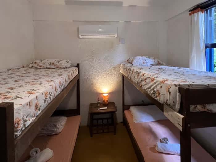 Dois beliches de madeira frente a frente, colchas de estampa floral em cima e lençóis rosa embaixo, com um criado-mudo e luminária acesa entre eles