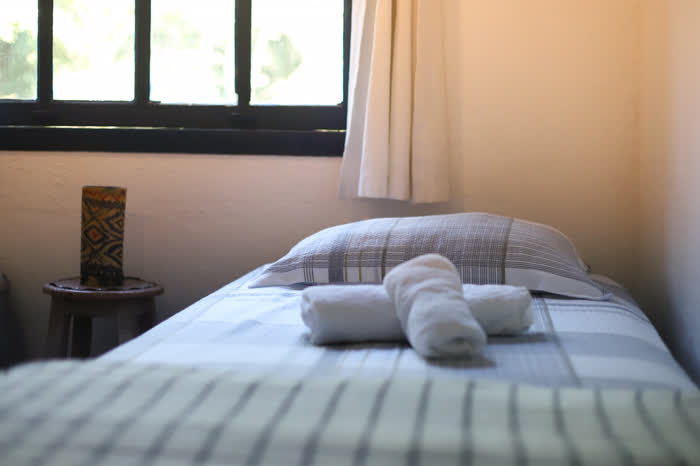 Cama de solteiro arrumada com toalhas enroladas sobre a colcha listrada, ao lado de um banquinho de madeira com luminária e a janela aberta para a mata