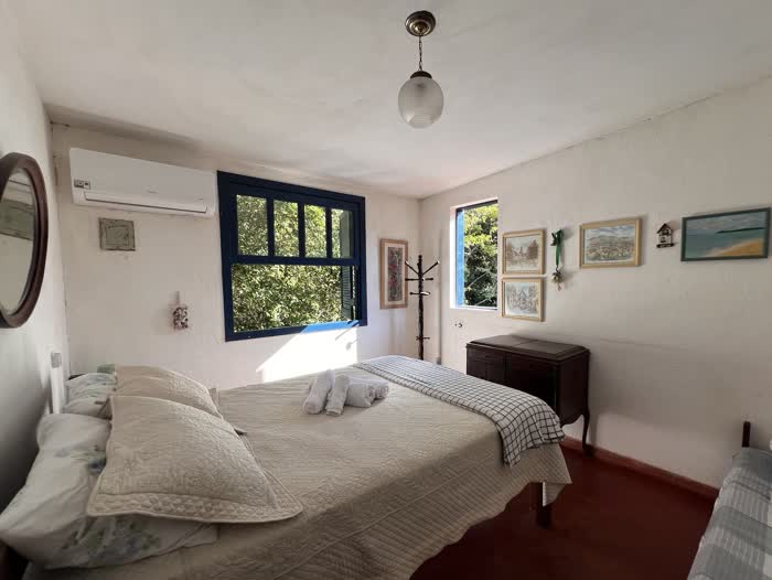 Quarto de casal com colcha branca, cabideiro de madeira, cômoda antiga e um conjunto de quadros de paisagem na parede