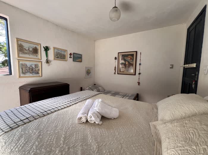 Cama de casal com colcha branca e toalhas enroladas, uma cama de solteiro ao lado e quadros emoldurados nas paredes brancas