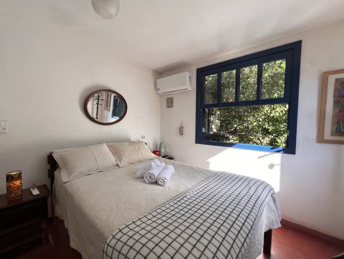 Quarto de casal com colcha branca matelassê, espelho oval na parede, ar-condicionado e a janela azul aberta para a folhagem