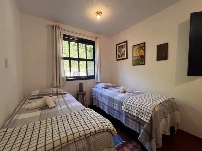 Quarto com duas camas de solteiro de colcha xadrez azul e manta, janela de cortina clara e três quadros pequenos na parede