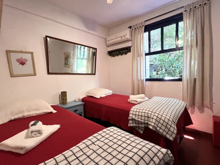 Quarto com duas camas de solteiro cobertas de colcha vermelha e manta xadrez, ar-condicionado e a janela aberta para a vegetação