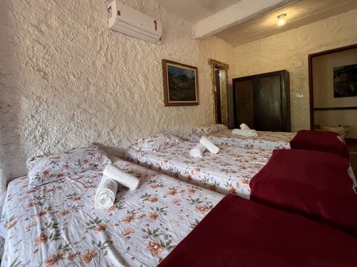 Quarto de parede rústica com três camas de solteiro forradas de colcha floral, toalhas enroladas em cada uma e um armário de madeira ao fundo