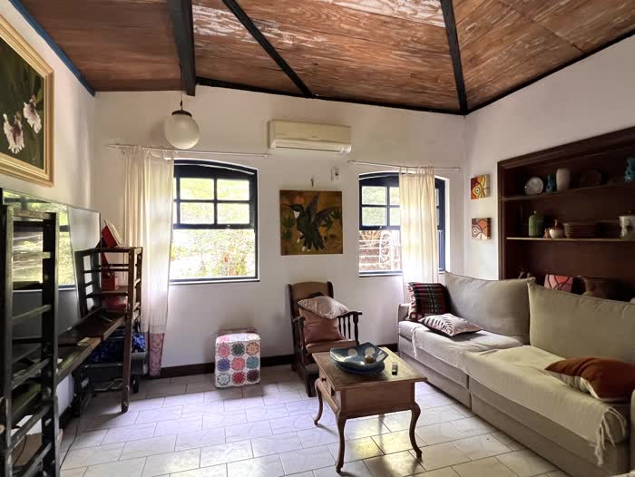 Sala de estar com teto de tábua corrida, sofá bege em L, poltrona de madeira e um quadro de beija-flor entre as duas janelas