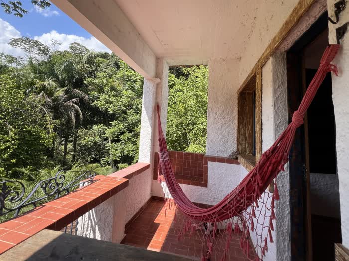 Varanda de piso de tijolo com uma rede vermelha armada, aberta para a mata e o céu azul