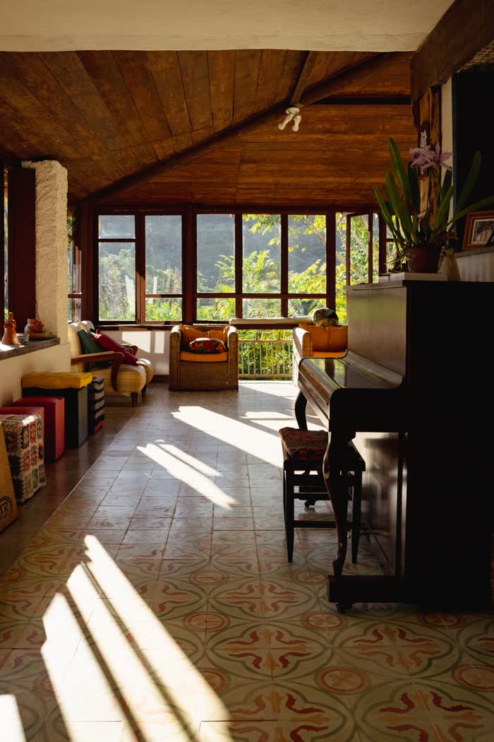 Sala da casa-sede no fim da tarde, com piano, poltronas de vime, uma orquídea florida sobre o piano e as janelas abertas para a mata